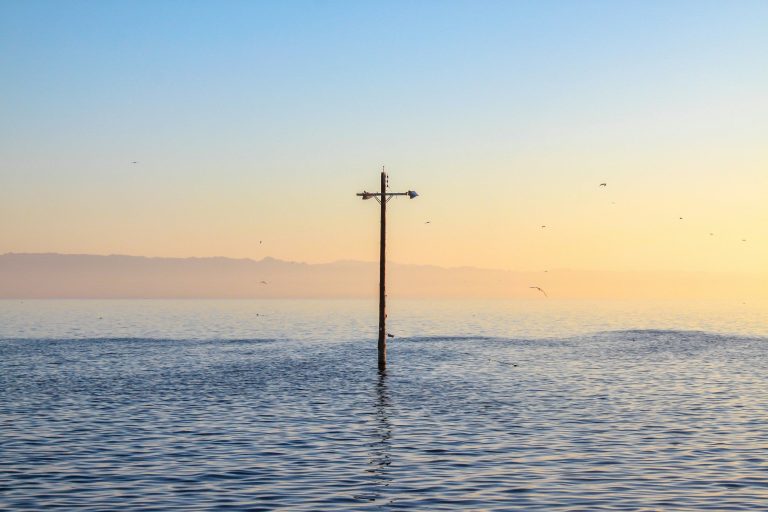 Überflutung Ein einzelnes Kreuz ragt aus ruhigem Wasser bei Sonnenaufgang.